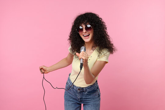 Beautiful young woman with microphone and sunglasses singing on pink background