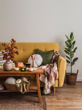 Autumn living room - yellow sofa with pillows and blankets, wooden oak bench with autumn flowers, pumpkins and lighted candles. Autumn mood interior