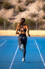 Beautiful tanned slim young runner girl, dressed in tight sportswear, running and sprinting on her back with her tail waving in the wind on a blue athletics track.