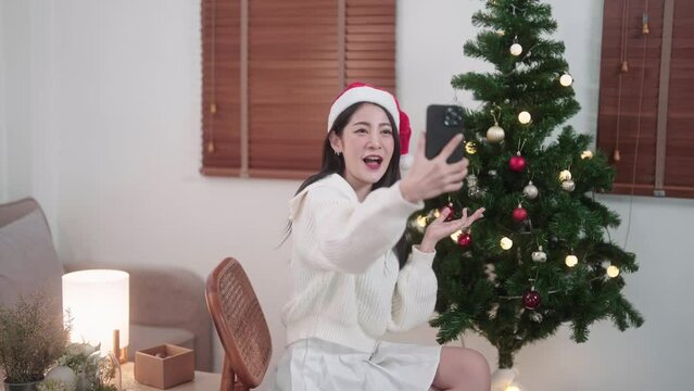 Young Asian Woman looking at camera doing vdo call or photographing illuminated christmas tree through smartphone at home, winter holiday concept.