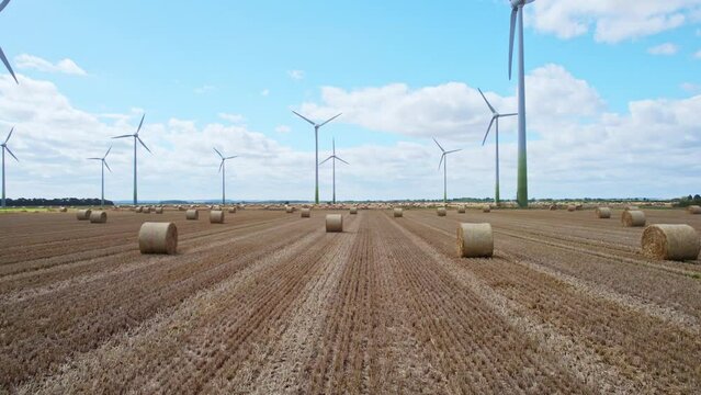 High in the sky, a captivating tableau unfolds as wind turbines spin within a Lincolnshire farmer's newly harvested field, where golden hay bales complete the picture.