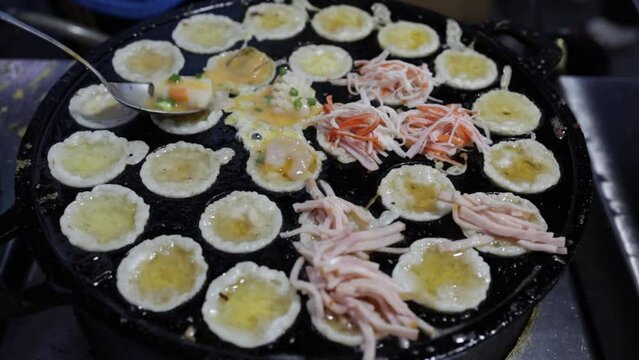 Street food chef putting seafood toppings on fried seafood mini pancakes in Thai-styled Takoyaki pan. Cooking of crispy fried seafood mini pancakes. Street food Thailand. Fried seafood omelet pancake.