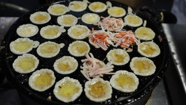 Food vendor putting ham on crispy fried mini pancakes using tongs in Thai-styled Takoyaki pan at market. Cooking crispy fried seafood mini pancakes. Street food Thailand. Fried seafood omelet pancake.