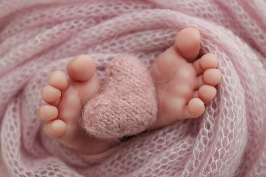 Closeup of toes, heels and feet of a newborn. Knitted pink heart in the legs of baby.The tiny foot of a newborn baby. Soft feet of a new born in a wool pink blanket. Macro studio photography. 