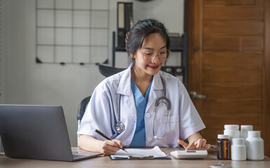 Portrait of cheerful smiling young female doctor in blue medical uniform typing on laptop computer, sitting at desk near window in modern office of medic clinic