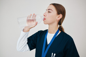Nurse, woman drinking water in bottle and health, wellness or body nutrition in studio isolated on white background in hospital. Medical professional, hydration and liquid of thirsty surgeon on break