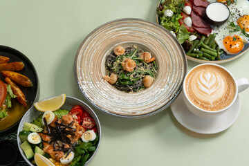 group photo of a perfect morning meal on a green background: latte coffee, green bowl with shrimps, fried eggs with toast, ham, green salad