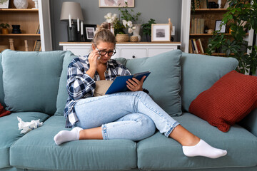 Sad woman crying while she reading a good paper book sitting on a sofa at home. Upset girl read love story cry of sadness - obrazy, fototapety, plakaty
