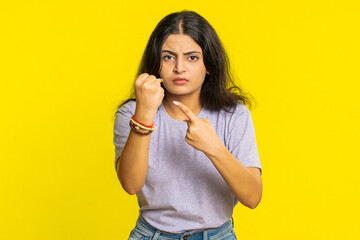 Aggressive angry Indian young woman trying to fight at camera, shaking fist, boxing with expression, punishment, threaten, bullying, abuse, mad fury. Arabian girl isolated on yellow studio background