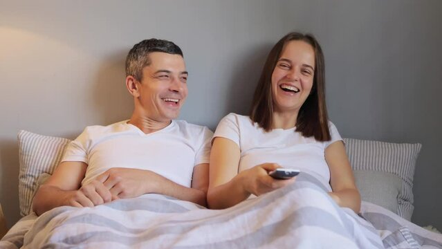 Joyful positive married couple lying in bed together and watching comedian movie laughing happily enjoying cozy lazy evening at home woman holding remote control for changing channels.