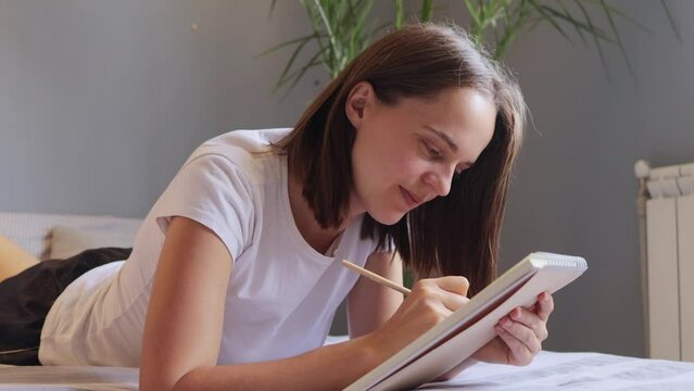 Attractive happy positive brown haired woman dressed in pajama makes notes in diary lies on bed answering letter focused on her writing enjoying pastime.