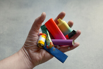 A white male holds a selection of discarded and broken electronic cigarette vapes, with their parts and batteries. Shot in front of a pale grey background.