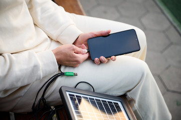 Pretty woman sitting on the bench and charging her phone with a portable solar panel. Solar battery...