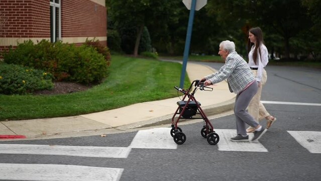 Side view of grandmother using a walker and granddaughter smiling and walking across crosswalk together outside.