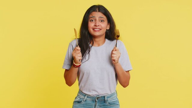 Ready to eat. Hungry Indian woman waiting for serving dinner dishes with with restlessness holding cutlery fork knife will appreciate delicious restaurant meal. Excited girl on yellow background