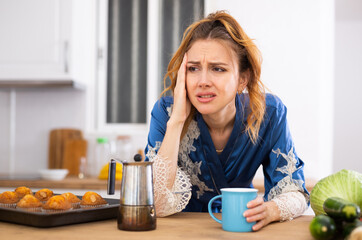 Young woman suffering from migraine, drinking coffee in home kitchen