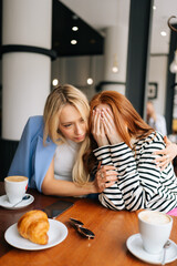 Vertical portrait of two best girlfriends sitting at indoors cafe and talking. Blonde female stroking hand, hugging, trying calm unhappy redhead friend, who cover face with hands and tearing.