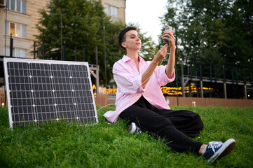 Fototapeta premium Young woman charging her phone by solar panel. Pretty female in casual clothes sitting on grass and pluging charger in her phone. Renewable energy concept.