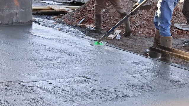 There is worker holding a steel fresno concrete trowel against freshly poured concrete as he smooths the plastering of new sidewalks in construction site