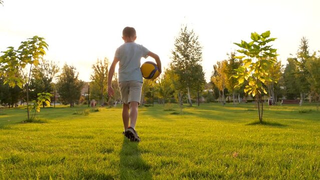 A child goes on the green grass holding a ball in his hands. The concept of a kids dream, sports, forward. Silhouette of a child at sunset with a ball. active children's games in the city park
