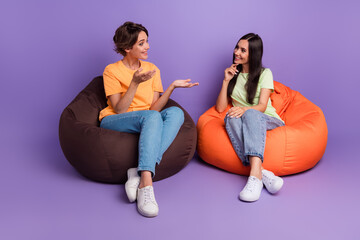 Photo of cheerful positive ladies wear t-shirts sitting bean bags talking isolated violet color background