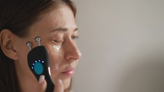 Female does microcurrent procedure for skin lifting. Brunette lady makes massage movements on face skin near eyes using microcurrent in bathroom