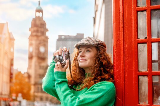 Young redhead woman in English style cap portrait close to red telephone box in English city taking  photos with vintage camera.  Old city  in the background.