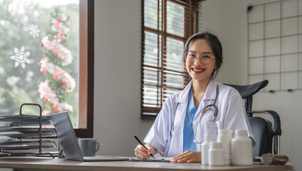 Portrait of cheerful smiling young female doctor in blue medical uniform typing on laptop computer, sitting at desk near window in modern office of medic clinic