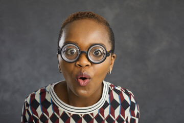 Close up portrait of funny shocked african american woman in glasses with magnifying glass. Woman with short hair and in a sweater on a gray concrete background. Concept of one's own vision.