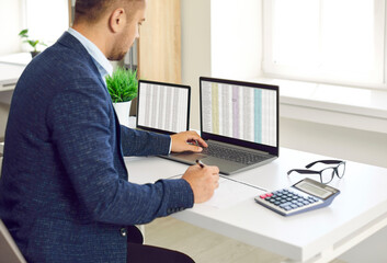 Busy concentrated male financier working in office with Excel spreadsheets and calculator. Man sits in front of two laptops, fills out paper documents and enters data into electronic files.