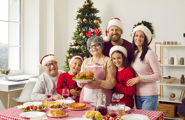 Cheerful family enjoying festive Christmas lunch meal at home. Portrait of happy grandmother in funny headband and apron holding roast turkey surrounded by the rest of the family in Xmas Santa caps