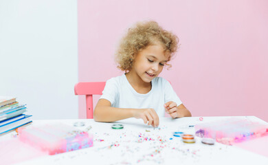 Cute little blonde girl making bead jewelry at a table in the room