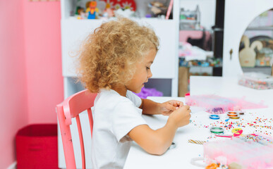 Cute little blonde girl making bead jewelry at a table in the room