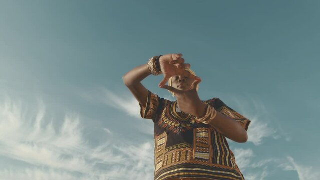 Low angle zoom out portrait of beautiful young African American woman in traditional ornate clothing making eye shape with hands while standing under clear blue sky on sunny day
