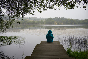 Rear view of silhouette of woman with blue raincoat sitting on the wooden pier in front of the lake...