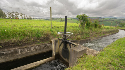 View of a metal gate of an irrigation canal with a background of planted fields on a cloudy day