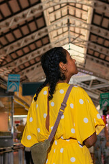 A young Latina, dressed in a vibrant yellow polka dot dress, enters a bustling market with her back to the camera