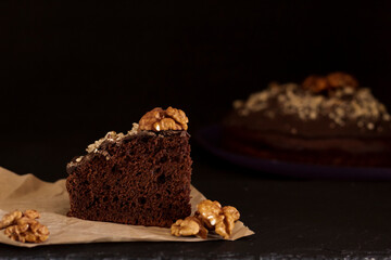 A piece of birthday cake sprinkled with walnuts. Close-up, front view. Brown pie on paper and black background.