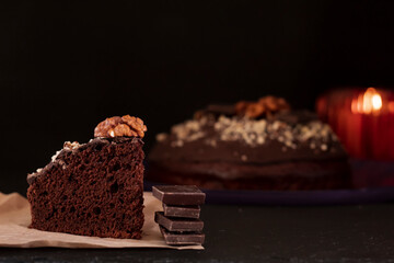 A piece of chocolate cake in the foreground is sprinkled with crushed walnuts on a dark background....