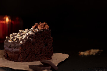 A piece of chocolate cake on paper with crushed nuts and frosting. Black background. Blurred bokeh background.