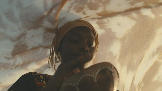 Low angle medium closeup of smiling young African American woman in yellow headscarf and ornate blouse playing djembe while standing under soft flowing fabric