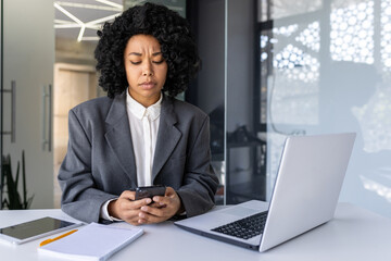 Upset young African American woman working in the office on a laptop, sitting at the desk, holding the phone and looking sadly at the screen.