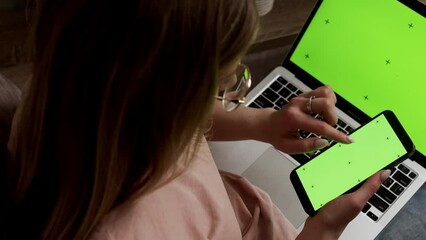 A woman uses a green smartphone screen with a Chromakey laptop. A close-up shot of a woman holding a cell phone while swiping her finger across a green screen and working with a laptop.