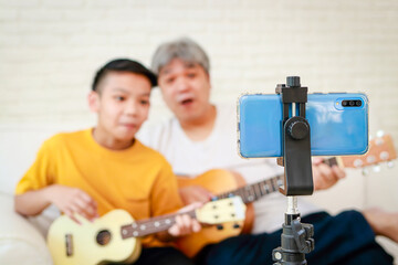 Happy Asian father and son playing music together Show live online on social media via smartphone. Studying music. Concert. Band