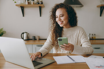 Young housewife woman wears casual clothes sweater work on laptop pc computer study use mobile cell...