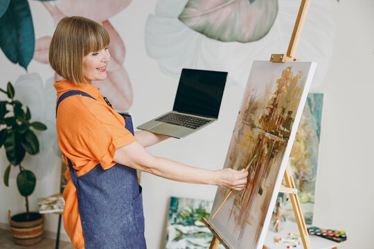Elderly artist woman 50 years old wears casual clothes stand near easel with painting artwork hold blank screen laptop pc computer spend free spare time in living room indoor. Leisure hobby concept.