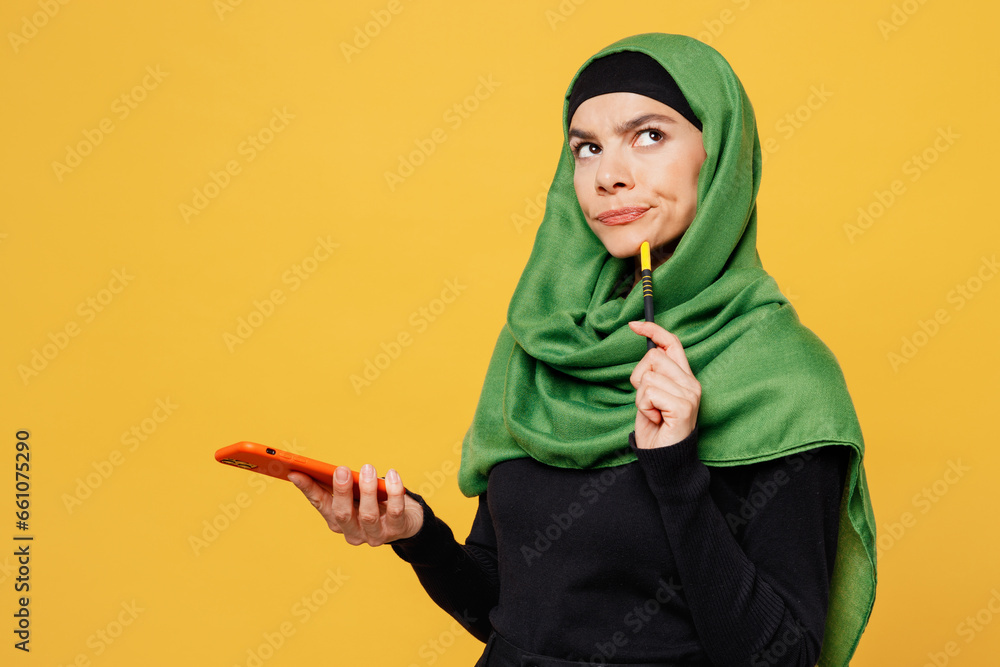 Wall mural Side view young minded arabian asian muslim woman wear green hijab abaya black clothes hold use mobile cell phone isolated on plain yellow background People uae middle eastern islam religious concept