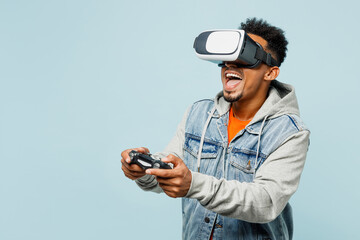 Young man of African American ethnicity in denim jacket orange t-shirt hold in hand play pc game with joystick console watching in vr headset pc gadget isolated on plain pastel light blue background - obrazy, fototapety, plakaty