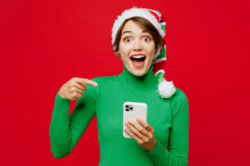 Young fun woman wear warm cozy green turtleneck Santa hat posing hold use point finger on mobile...