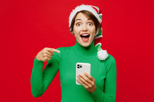 Young fun woman wear warm cozy green turtleneck Santa hat posing hold use point finger on mobile cell phone isolated on plain red background. Happy New Year 2024 celebration Christmas holiday concept.